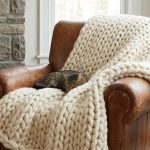 A tabby cat sleeps on a chunky cream knit blanket draped over a brown leather armchair in a cozy living room